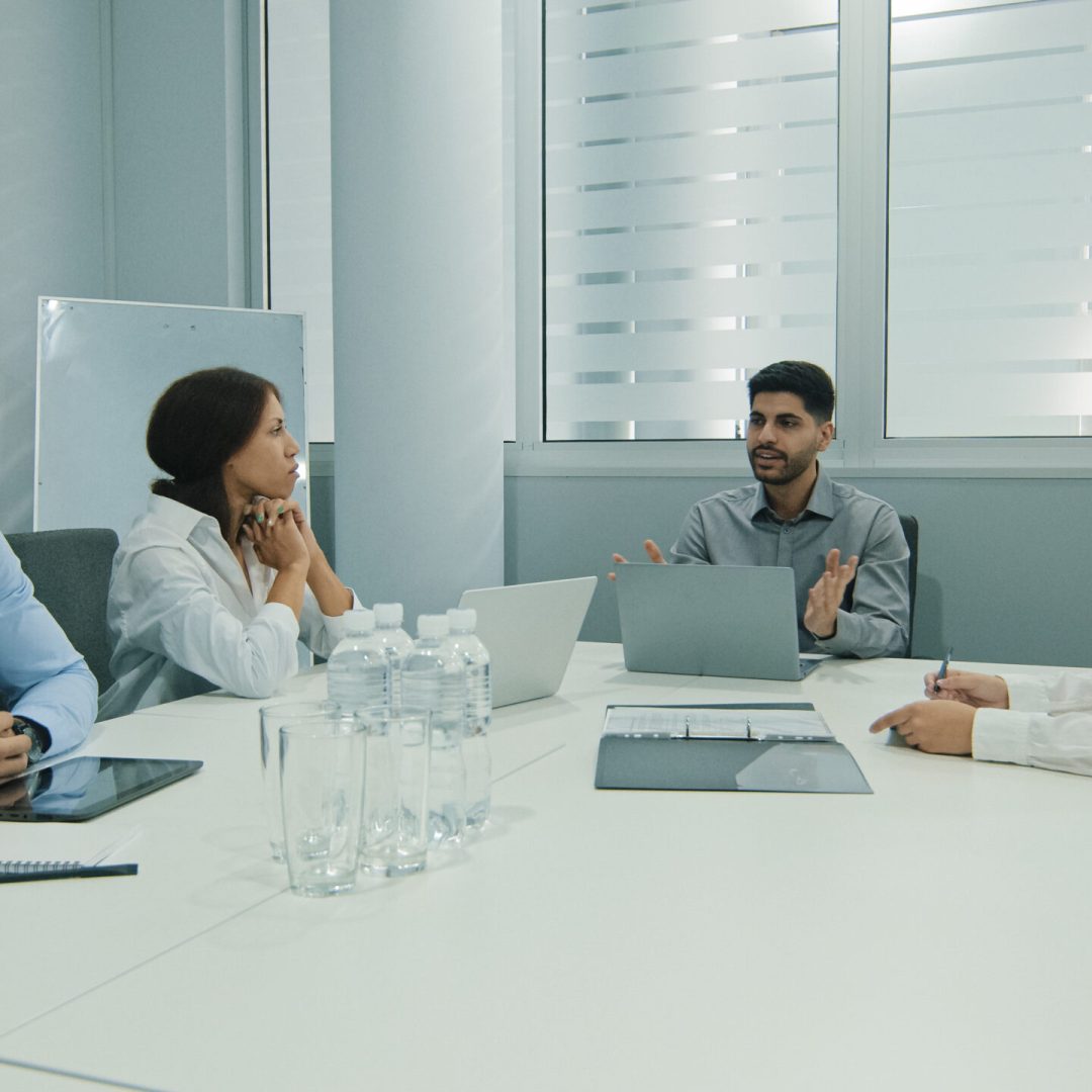 Young indian team leader sitting with colleagues, discussing issues at negotiations meeting. Skilled millennial diverse employees working together at office developing startup marketing strategy. High quality photo